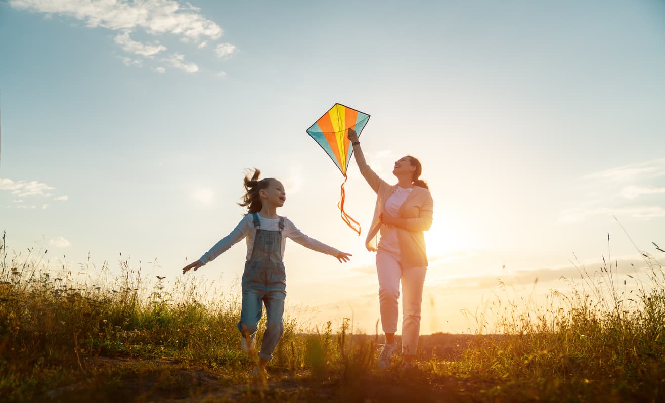 Woman and child running through a field with a multi-colored kite at sunset.