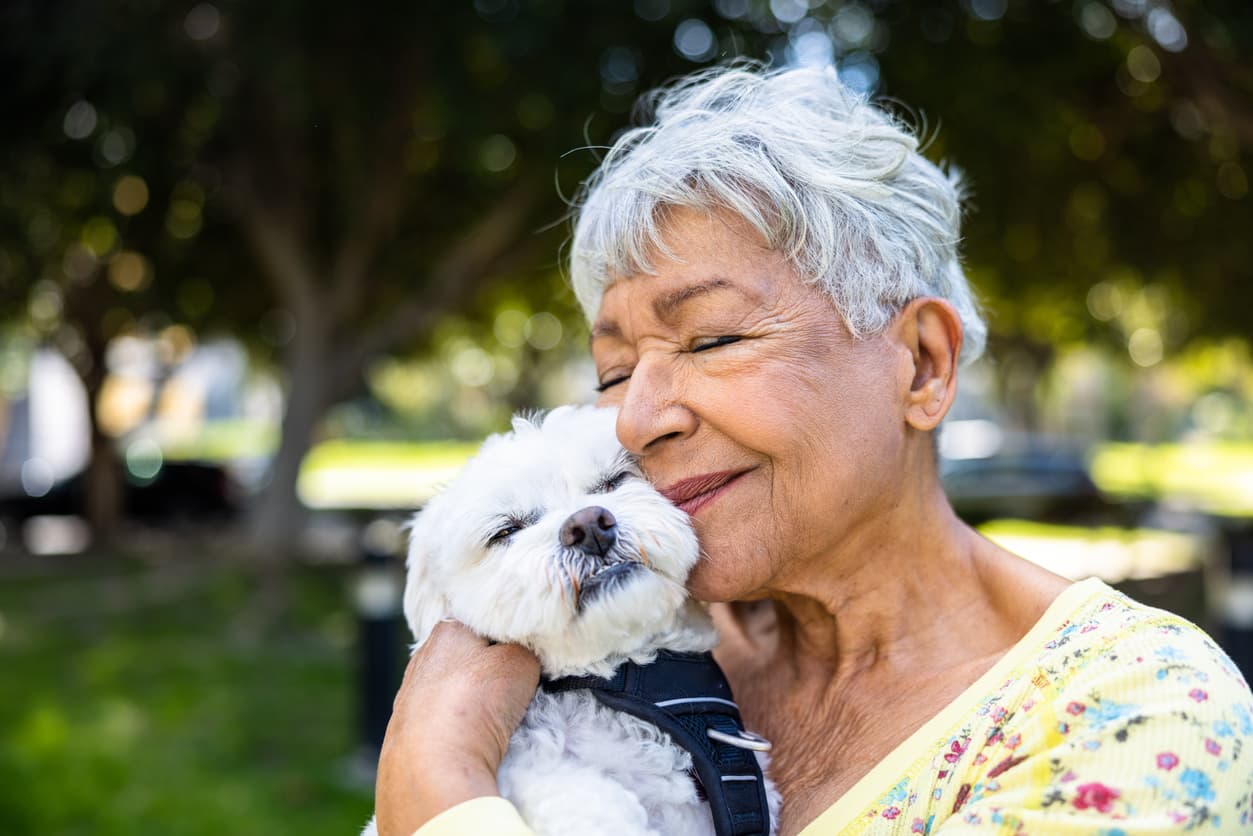 Happy elderly woman with short white hair holding small white dog to her face.