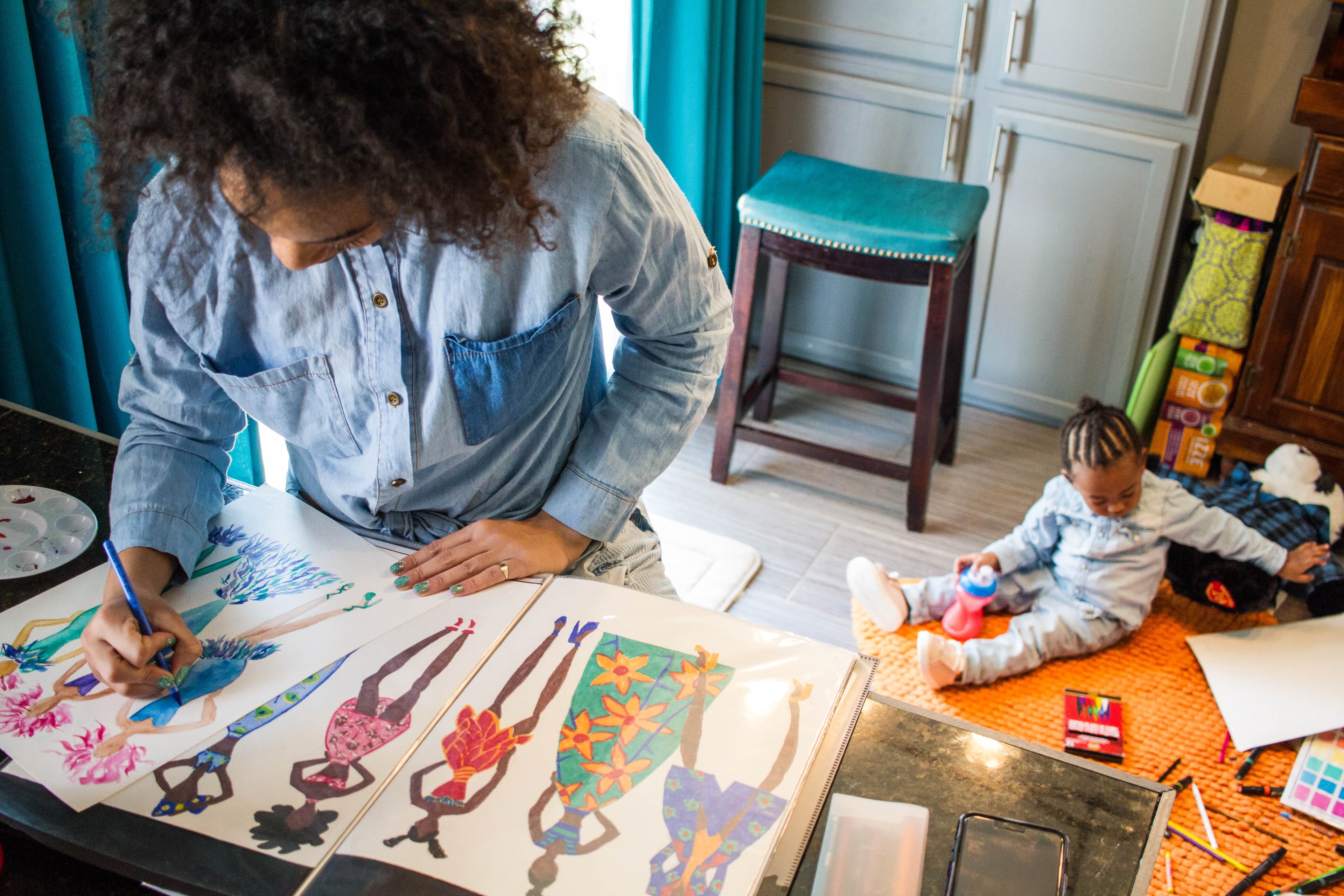 Woman standing at a desk in a bright home workspace drawing colorful fashion designs while a toddler plays with toys on the floor nearby.